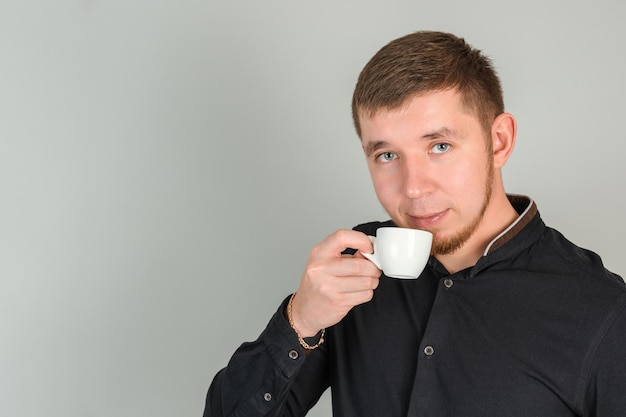 A young bearded red-haired man drinks coffee from a small white mug looking at the camera, horizontal photo