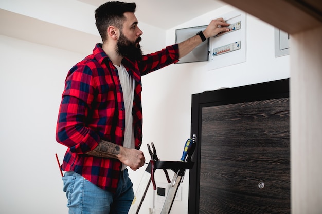 Young bearded repairman standing on ladder and repairing fuse box.