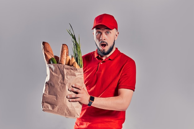 Young bearded shocked surprised delivery man in red uniform holds package with bread and vegetables isolated on gray background.