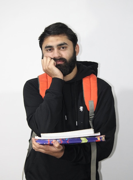 Фото Молодой бородатый студент в черном капюшоне с книгами, стрессовый и нервный у белой стены 2