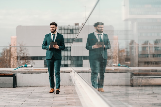 Young bearded successful businessman in formal wear walking on roof and holding tablet.