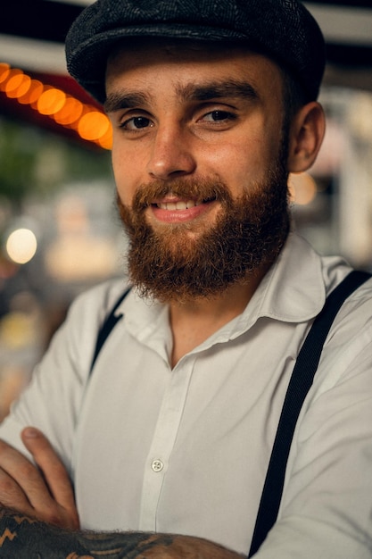 Young bearded tattooed man in a cafe on the street with a glass of wine. A romantic guy in a white cap and suspenders in the city. Peaky Blinders. old fashionable retro.