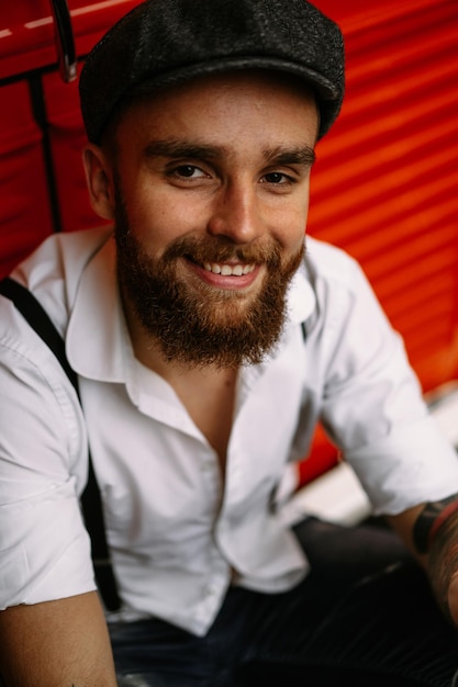 Young bearded tattooed man portrait close-up. A romantic guy in a white shirt, cap and suspenders. Peaky Blinders. old-fashioned, retro.