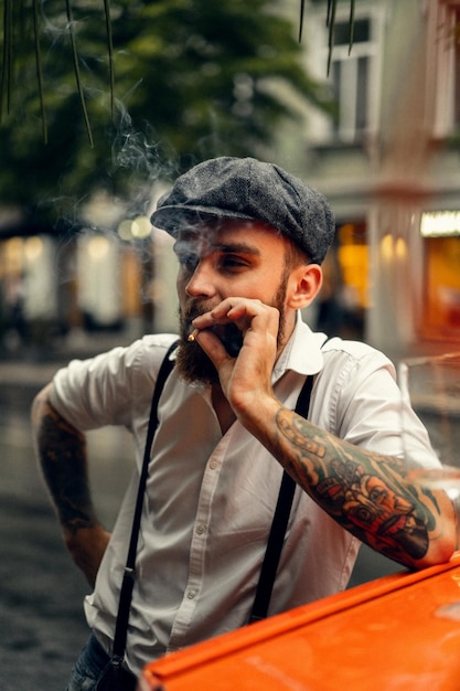 Young bearded tattoord man in a cafe on the street smokes a cigarette Romantic guy in a white shirt cap and suspenders in the city Peaky Blinders old fashionable retro