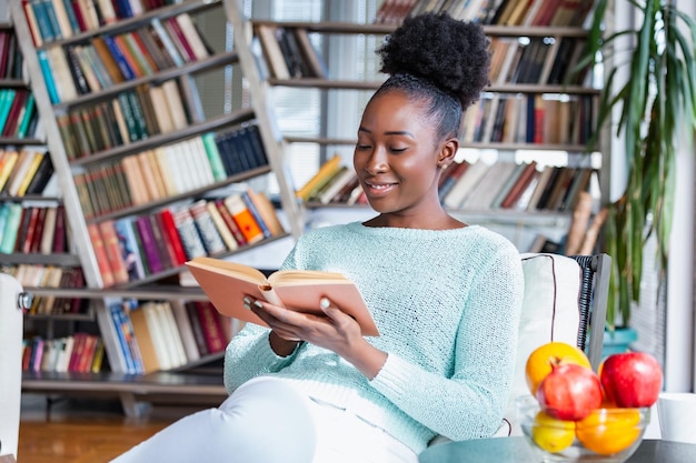 Photo young beautiful african american girl reading a book on the couch with the library bookshelves in the back beautiful woman on a white sofa reading a book