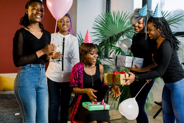 Young beautiful African American girls with balloons and hats celebrate birthday and give birthday gifts.