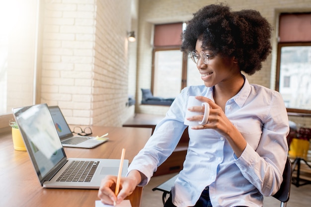 Молодая красивая африканская бизнесвумен пьет кофе, сидя с ноутбуком в кафе или офисе