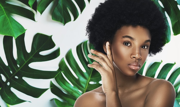 熱帯の葉に完璧な滑らかな肌を持つ若くて美しいアフリカの女性。自然派化粧品とスキンケアのコンセプト。