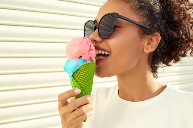 A young beautiful africanamerican girl in a white tshirt and light jeans eats ice cream against a light wall on a sunny day