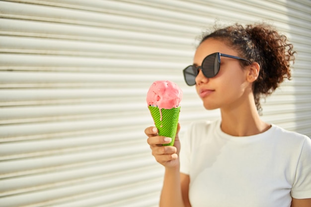 흰색 티셔츠와 가벼운 청바지를 입은 한 젊은 아프리카계 미국인 소녀가 작은 초점 영역에 초점을 맞춘 화창한 날 아이스크림을 먹습니다