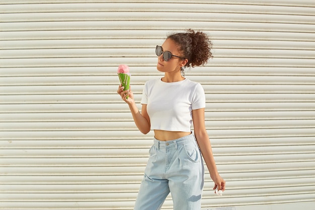 A young beautiful AfricanAmerican girl in a white tshirt and light jeans eats ice cream on a Sunny day selective focusing small focus area