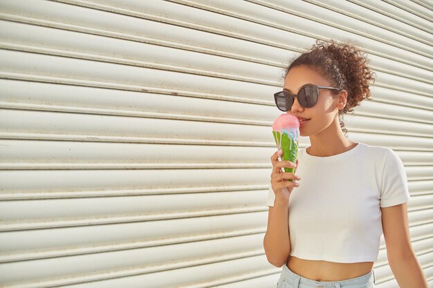 Молодая красивая афроамериканка в белой футболке и светлых джинсах ест мороженое в солнечный день, избирательно фокусируясь на небольшой области фокусировки