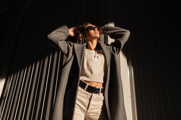 Фото Молодая красивая и стильная модель девушка в солнечных очках в модном винтажном осеннем пальто с вязанным свитером и брюками позирует на улице возле бетонной стены при солнечном свете