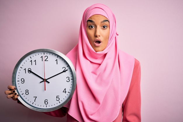 Giovane bella donna araba che indossa l'hijab musulmano rosa che fa il conto alla rovescia tenendo un grande orologio spaventato dallo shock con una faccia a sorpresa impaurita ed eccitata con l'espressione di paura