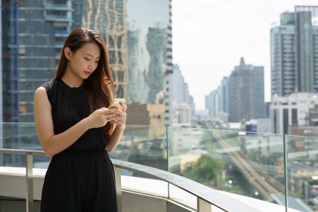 Young beautiful Asian businesswoman using phone against view of the city