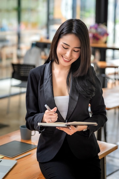 Молодая красивая азиатская женщина-бизнесвумен, сидящая на краю офисного стола, держа ручку и документ.