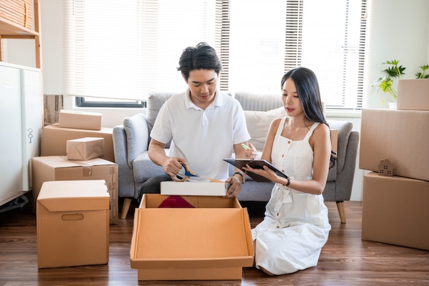 Young beautiful asian couple in love moving to new home, sitting on the floor very happy and cheerful for new apartment around cardboard boxes and holding cardboard boxes while moving home