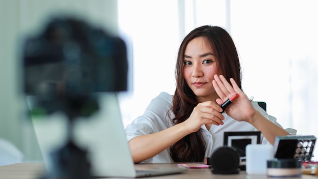 Молодая и красивая азиатская девушка показывает помаду на камеру и сравнивает с цветом кожи во время трансляции или записи видео об обзоре косметики и бьюти-блогере. Интернет-продажи и концепция маркетинга.