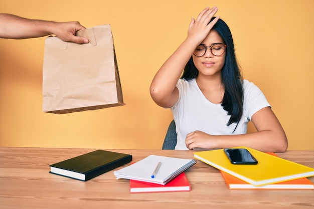 Young beautiful asian girl sitting on the table stuying getting take away food surprised with hand on head for mistake remember error forgot bad memory concept