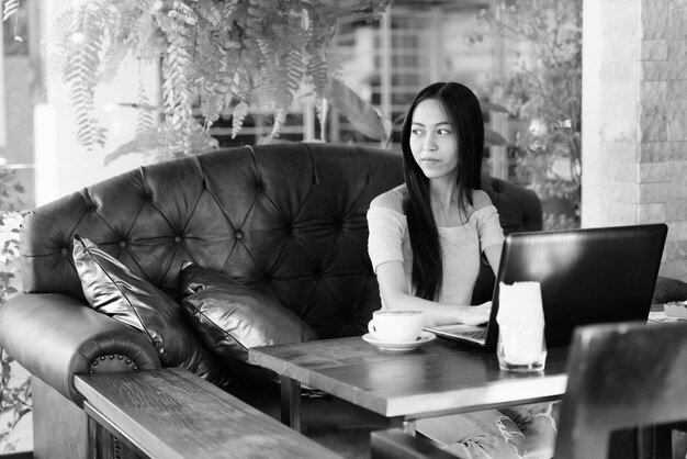 Young beautiful Asian girl sitting while thinking at outdoor coffee shop with laptop on wooden table