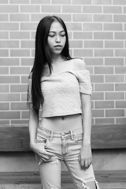 Young beautiful Asian girl standing while thinking and looking down on brick wall