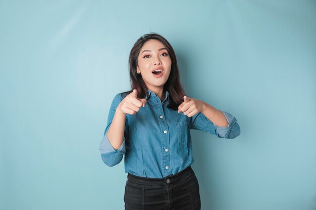 Young beautiful Asian girl wearing a casual blue shirt standing over isolated blue background pointing fingers at the camera with a happy and funny face Good energy and vibes