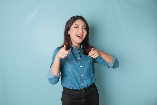 Young beautiful Asian girl wearing a casual blue shirt standing over isolated blue background pointing fingers at the camera with a happy and funny face Good energy and vibes