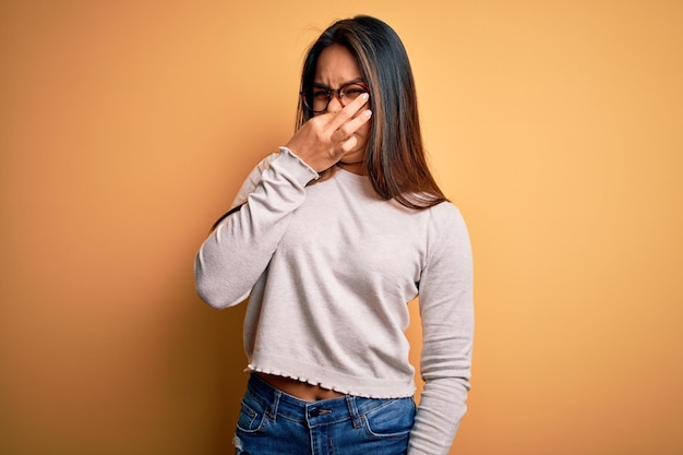 黄色の背景にカジュアルなセーターと眼鏡をかけた若い美しいアジアの女の子が何か臭くて嫌な耐え難い臭いを嗅ぎ、指を鼻に当てて息を止める 悪臭