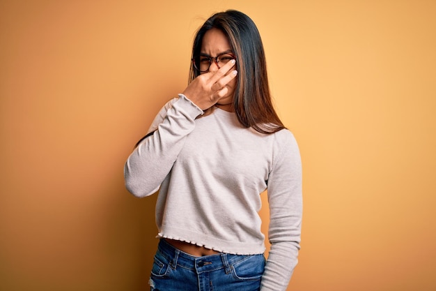 Foto giovane bella ragazza asiatica che indossa maglione casual e occhiali su sfondo giallo annusare qualcosa di puzzolente e disgustoso odore intollerabile trattenere il respiro con le dita sul naso cattivo odore