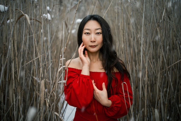 Photo young beautiful asian girl with natural make up in red silk dress in winter outdoor with dry reeds