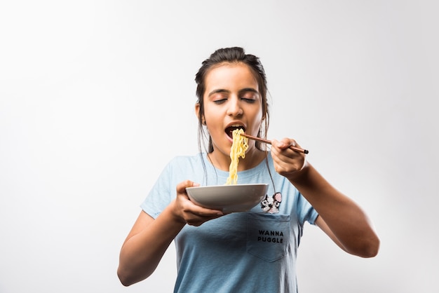 箸を使ってインスタントラーメンを食べる若い美しいアジアのインドの女の子。白い背景の上に孤立して立っています。セレクティブフォーカス