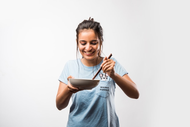 Молодая красивая азиатская индийская девушка ест лапшу быстрого приготовления, используя палочки для еды. стоя, изолированные на белом фоне. выборочный фокус