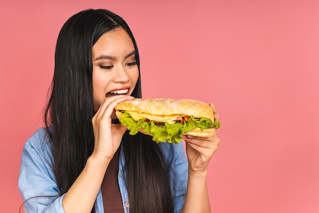 Young beautiful asian japanese chinese woman eating sandwich or big burger with satisfaction Girl enjoys tasty hamburger takeaway diet concept standing isolated over pink background