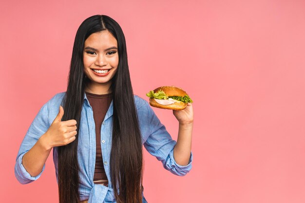 Giovane bella donna cinese giapponese asiatica che mangia panino o hamburger grande con soddisfazione la ragazza gode del concetto di dieta da asporto di hamburger gustoso in piedi isolato su sfondo rosa