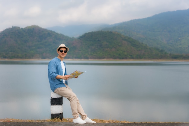 Young beautiful Asian man traveler with map in hands and wear sunglass looking and happy to see landscape view sits on a lake with beautiful mountain view in Thailand. Solo man travel
