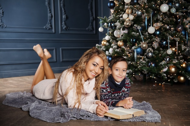 Young beautiful Asian mother with her little son sitting near a decorated Christmas tree and reading a book. 