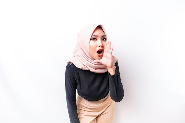 Young beautiful Asian Muslim woman wearing a hijab shouting and screaming loud with a hand on her mouth communication concept