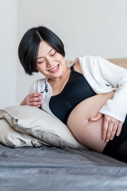 Giovane bella donna incinta asiatica che si trova a letto e tocca la sua pancia a casa