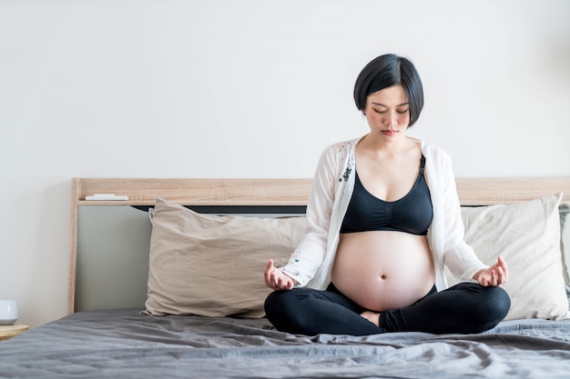 Young beautiful Asian pregnant woman meditating or doing yoga exercise at home. Relaxation yoga lotu