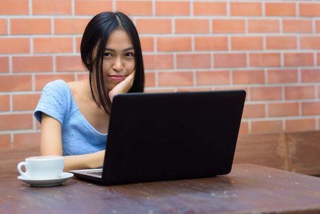 Young beautiful Asian teenage woman resting chin on hand with laptop and cappuccino on wooden table