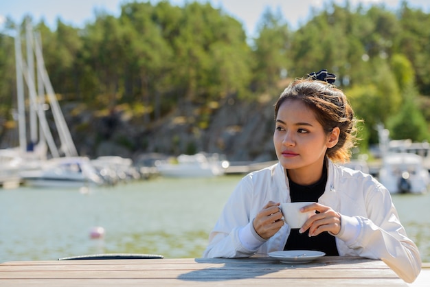 Giovane bella donna turistica asiatica che pensa mentre beve il caffè dal molo