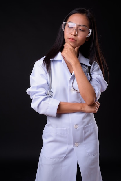  young beautiful Asian woman doctor as scientist with protective glasses against black wall