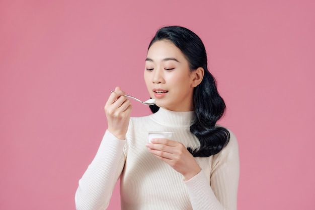 Young beautiful asian woman eating yogurt with spoon isolated pink background