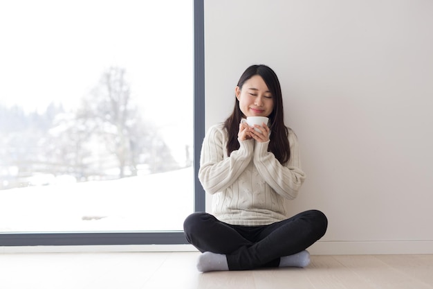 寒い冬の日に窓の近くの床で朝のコーヒーを楽しむ若い美しいアジアの女性