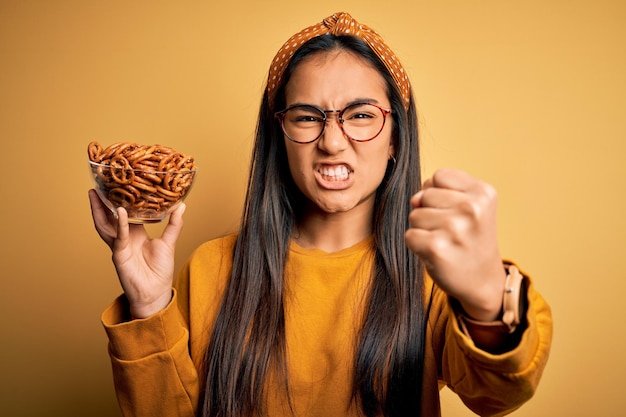 黄色の背景にドイツの焼きプレッツェルを入れたボウルを持つ若い美しいアジアの女性がイライラしてイライラして怒り狂って叫び、手を上げた怒りのコンセプトで叫ぶ