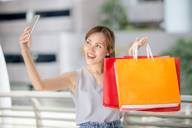 Young beautiful Asian woman holding her smartphone to taking a selfie with her shopping bags in the city street