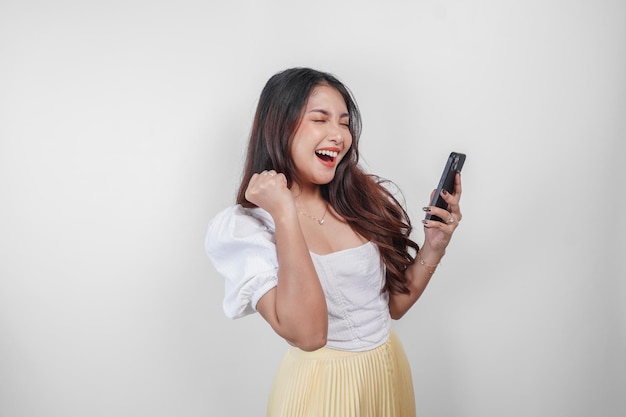 A young beautiful asian woman is cheering while holding her phone isolated by white background