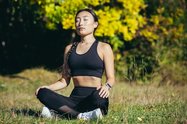 Young beautiful asian woman performs fitness yoga exercises, sitting in lotus position on the grass and meditating in the park