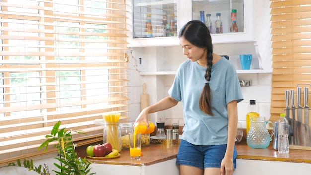 Young beautiful asian woman pouring orange juice on glass while standing in kitchen, Asian girl drinking orange juice for healthy, Teen woman and healthy lifestyle