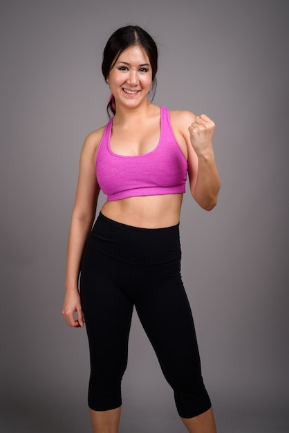 Young beautiful Asian woman ready for gym against gray wall